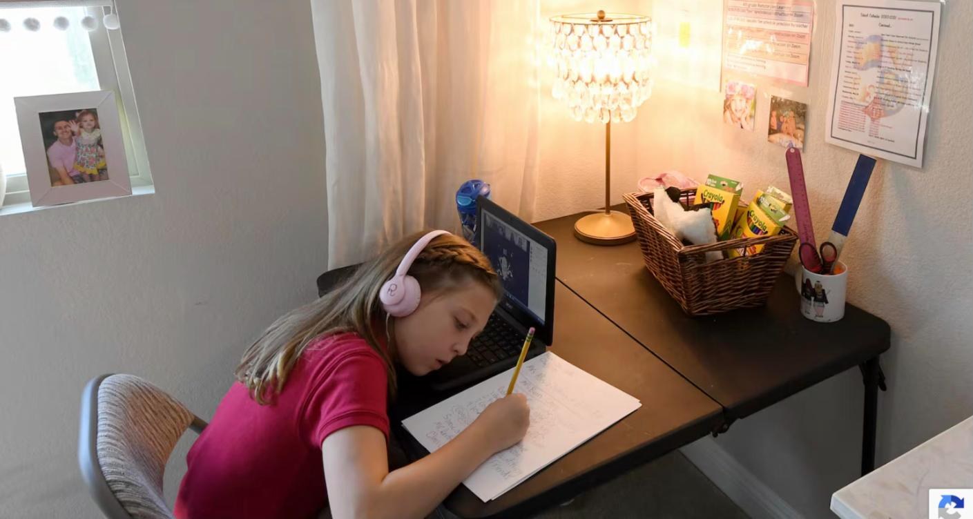 A young girl wearing headphones writes at her desk with a laptop