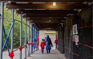A parent and child walk under sidewalk scaffolding outside a school
