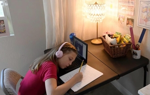 A young girl wearing headphones writes at her desk with a laptop